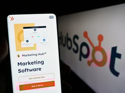 In the foreground, a smart phone screen is on HubSpot's Marketing Hub page. In the background, there is a blurry HubSpot booth skin, like what one would see at a conference.